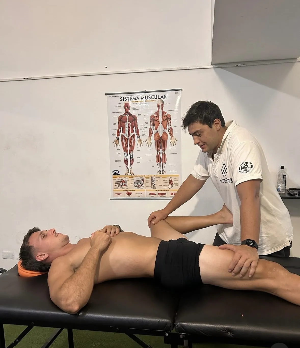 Bernardo Quaranta evaluando la cadera de un paciente, junto a un afiche del sistema muscular